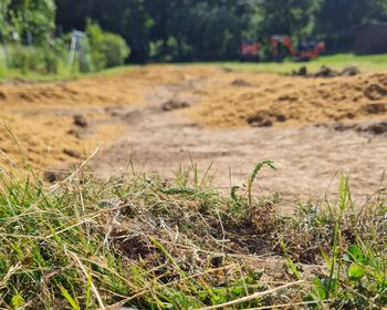 Vorne im Bild Gras und leicht angehäufter Sand, im Hintergrund eine landwirtschaftliche Maschine.
