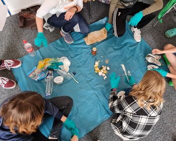 Kinder und Jugendliche mit blau behandschuhten Händen umringen eine blaue, auf dem Boden ausgebreitete Plastikfolie.