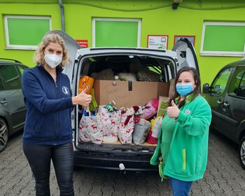 Zwei junge Frauen vor den Spenden im geöffneten Kofferraumes eines Kleintransporters mit "Daumen hoch" Zeichen.