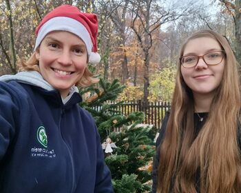 Zwei junge Frauen lächeln in die Kamera, eine davon trägt eine rot-weiße Weihnachtsmann-Mütze. Im Hintergrund ein grüner Weihnachtsbaum mit einem Hänger.