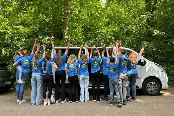 Ein Gruppe Kinder, Jugendlicher und Erwachsener steht mit dem Rücken der Kamera zugewandt vor einem weißen Kleinbus.