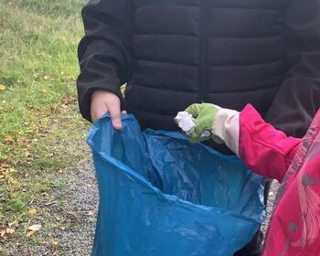 Ein Kind mit behandschuhten Händen wirft ein zerknülltes Papier in einen großen blauen Müllsack.