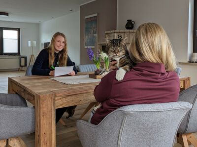 Zwei Frauen sitzen an einem Holztisch. Eine der beiden in Tierheim-Kleidung macht sich Notizen, der andere Frau, die von hinten sichtbar ist, schaut eine graugetigerte Katze über die Schulter.