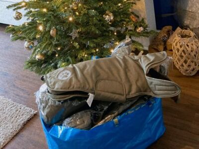 Eine große blaue Kunststofftasche, randvoll gefüllt mit neuen, teils verpackten Hundemänteln steht auf einem Holzfußboden vor einem festlich geschmückten Weihnachtsbaum.