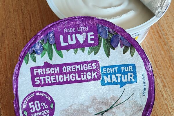 Geöffneter Becher des Luve-Streichglücks, daneben der Deckel mit Aufdruck in lila-blau-weißer Farbe.